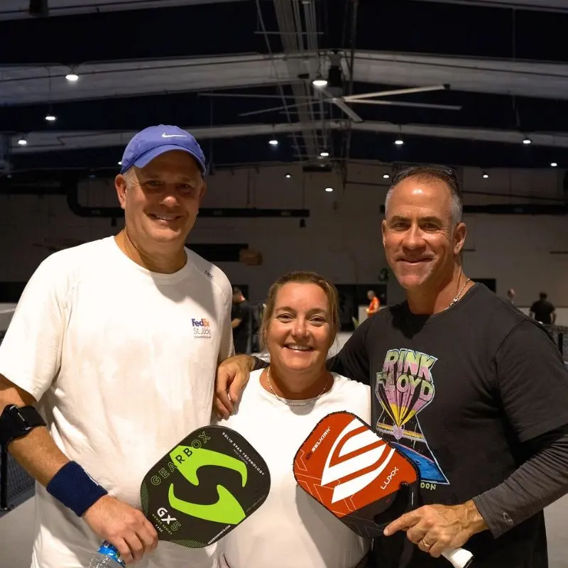 A coach and two players smiling on court with their paddles